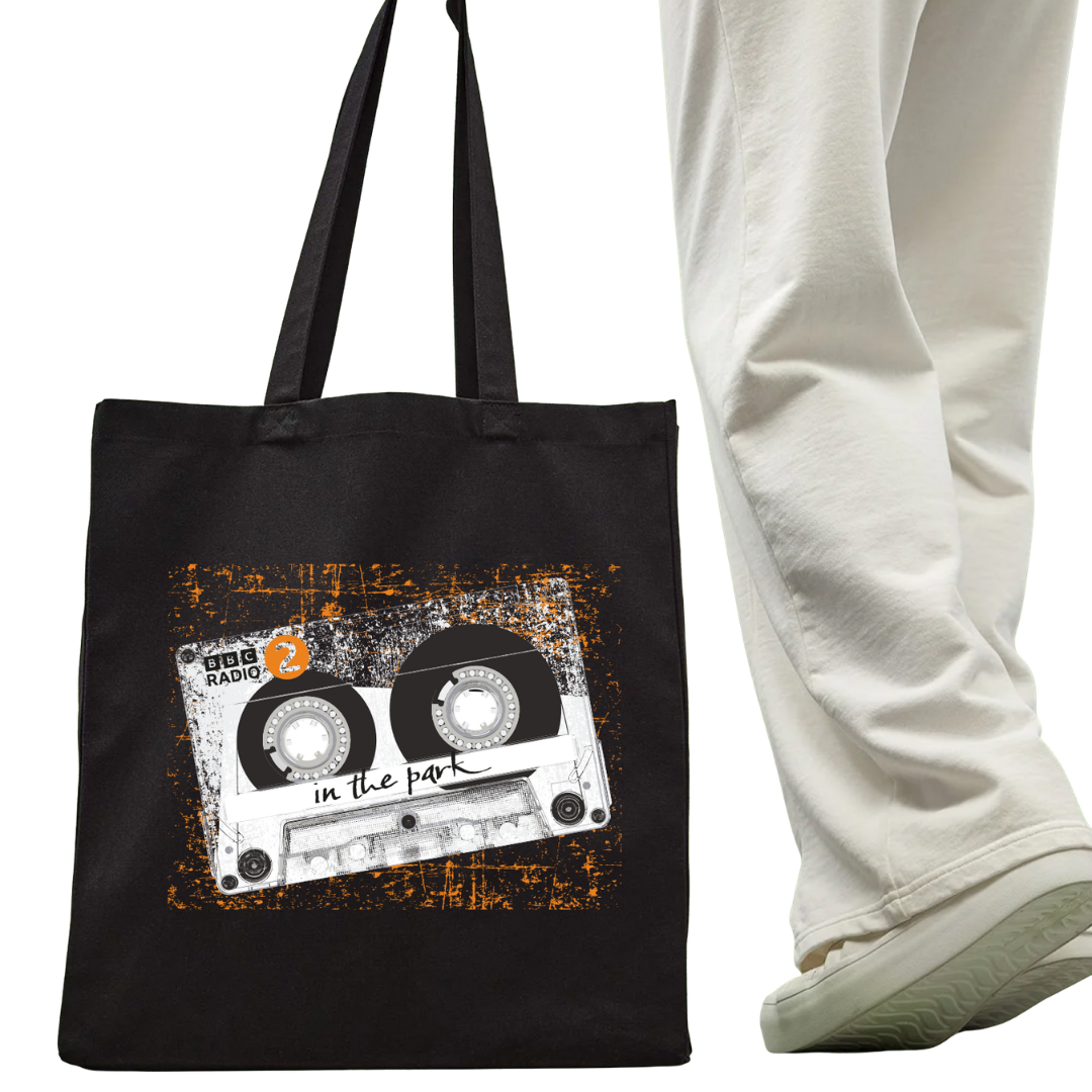 A person in white pants and sneakers stands next to the Click & Collect BBC Radio 2 in the Park XL Tote Bag from BBC Radio 2 Click & Collect 2026, which features a cassette tape design and “BBC Radio 2 in the park” in white text.