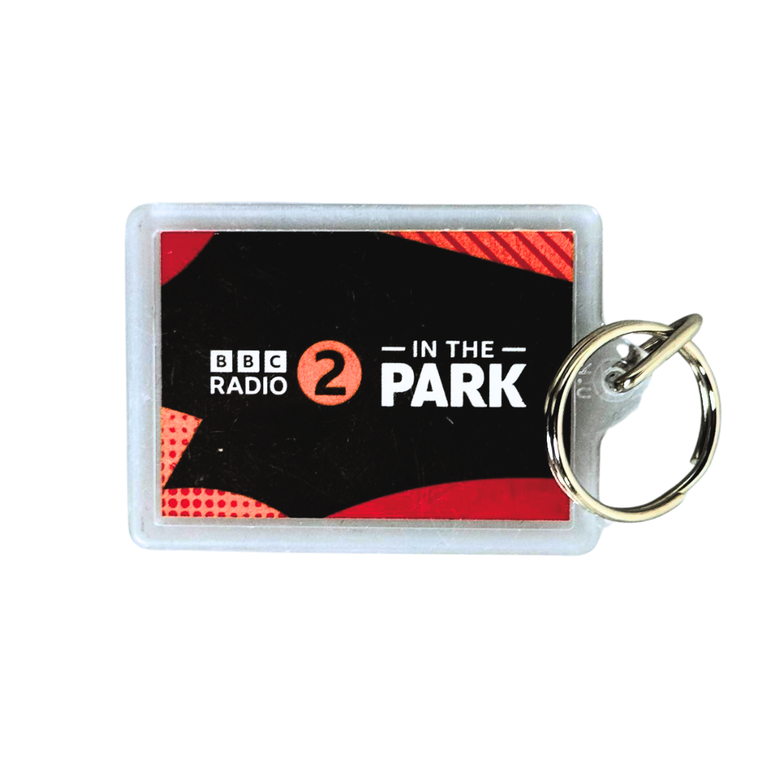 The BBC Radio 2 In The Park Keyring by Event Merch (National Merchandise) is a rectangular keyring with a metal ring, featuring a black and red design and "BBC Radio 2 In The Park" text.